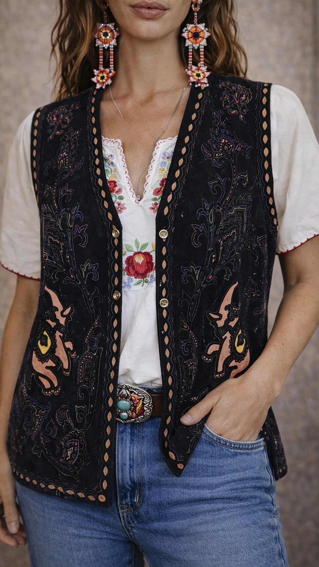Person wearing a black embroidered vest over a white blouse with floral design, paired with blue jeans.