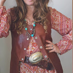 Woman wearing a brown hat, patterned dress, and leather vest with a belt buckle.