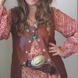Woman wearing a brown hat, patterned dress, and leather vest with a belt buckle.