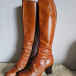Pair of brown leather knee-high boots on a light gray surface with a textured beige fabric background.
