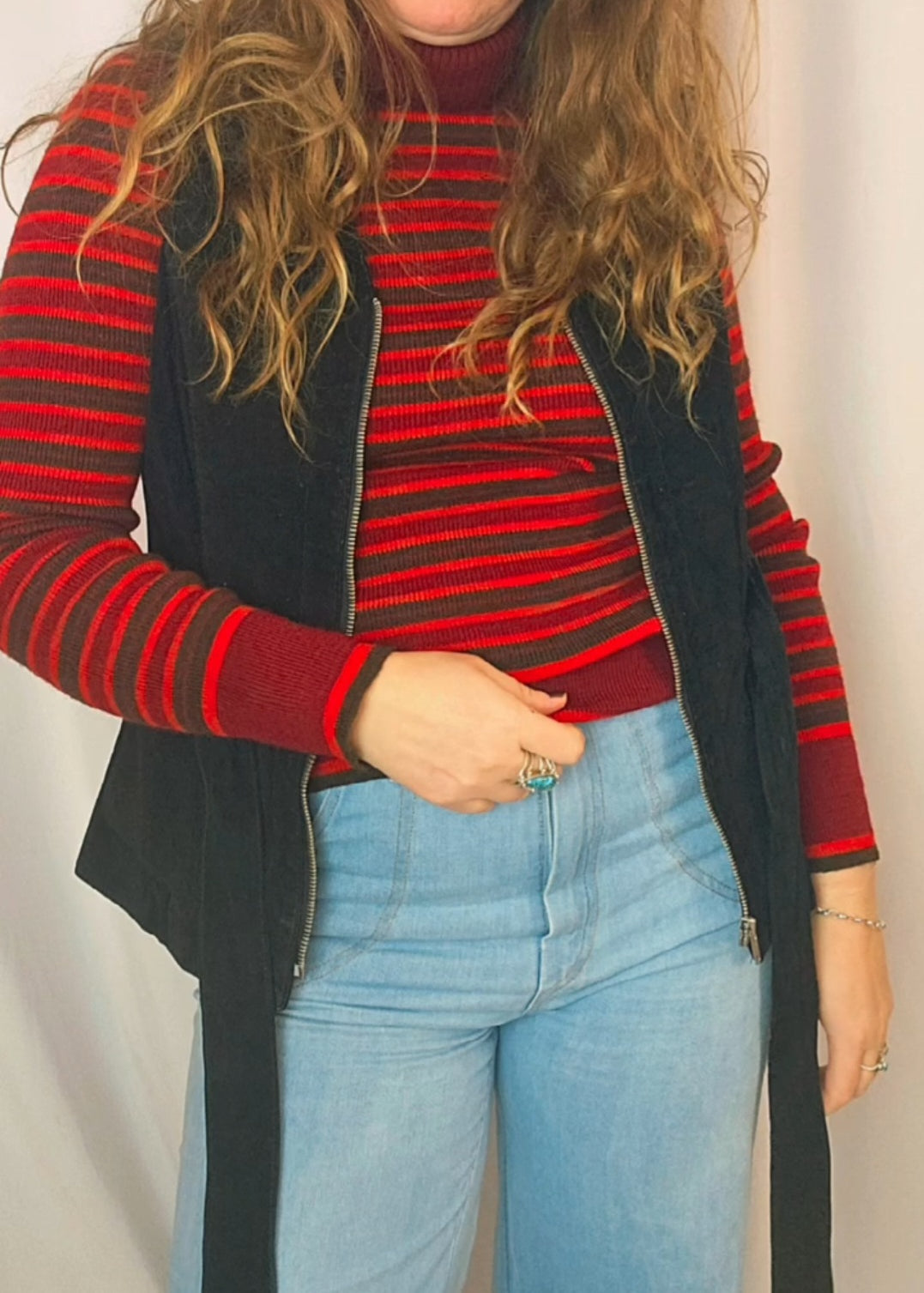 Person wearing a red and black striped sweater with a black vest and blue jeans against a plain background