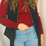 Person wearing a red and black striped sweater with a black vest and blue jeans against a plain background