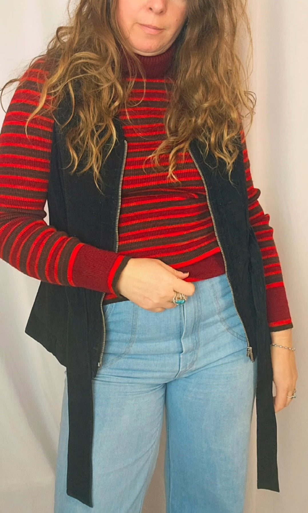 Person wearing a red and black striped sweater with a black vest and blue jeans against a plain background