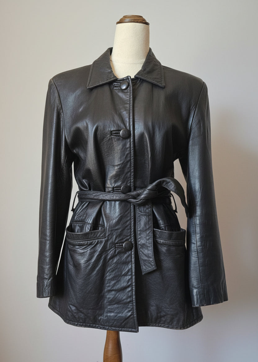 A black leather jacket with a matching tie belt, displayed on a mannequin torso. The jacket features a pointed collar, button closures, and front patch pockets, photographed against a neutral indoor background.