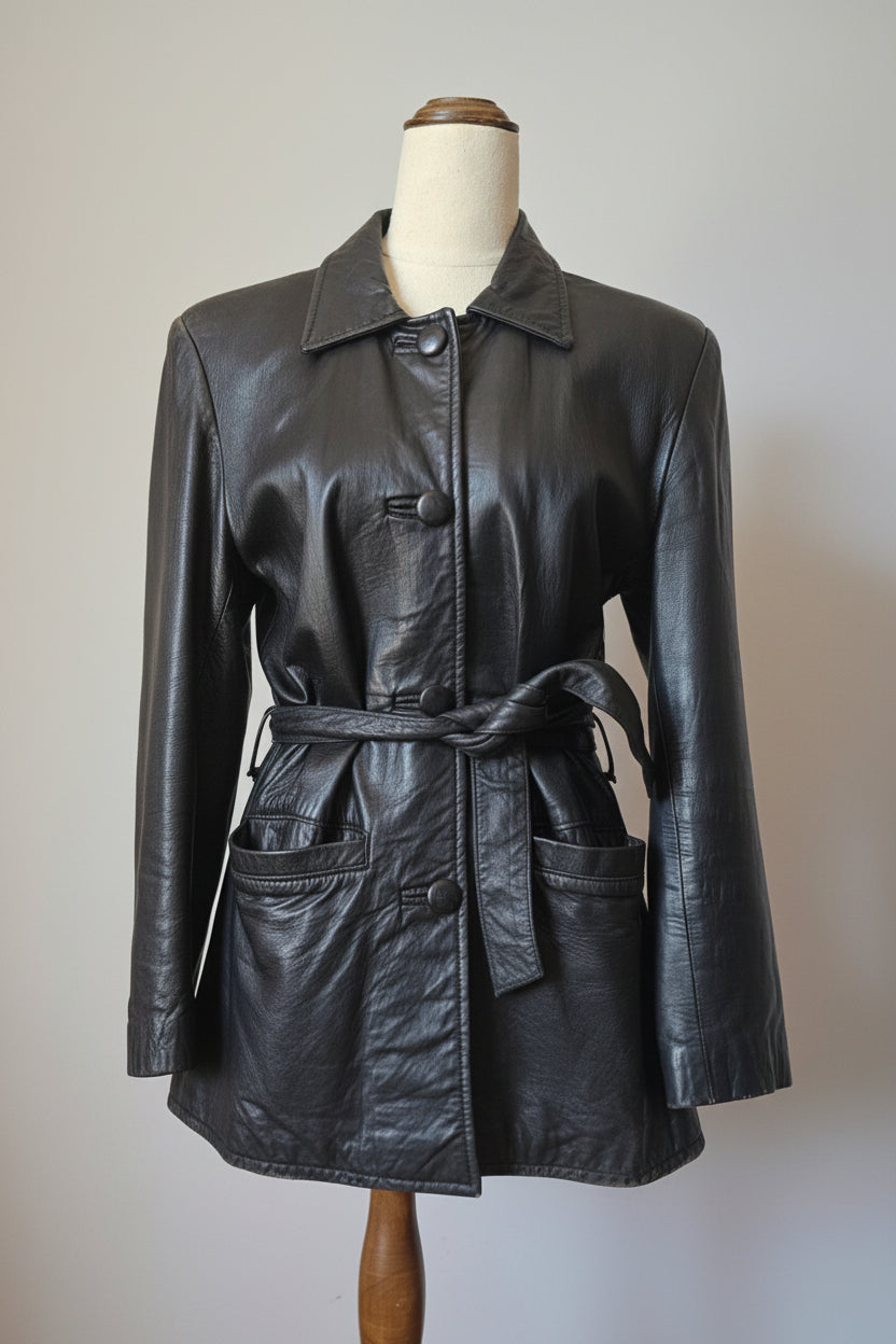 A black leather jacket with a matching tie belt, displayed on a mannequin torso. The jacket features a pointed collar, button closures, and front patch pockets, photographed against a neutral indoor background.