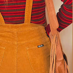 Person wearing mustard yellow overalls with a red and black striped shirt, holding a brown leather bag.