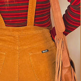 Person wearing mustard yellow overalls with a red and black striped shirt, holding a brown leather bag.