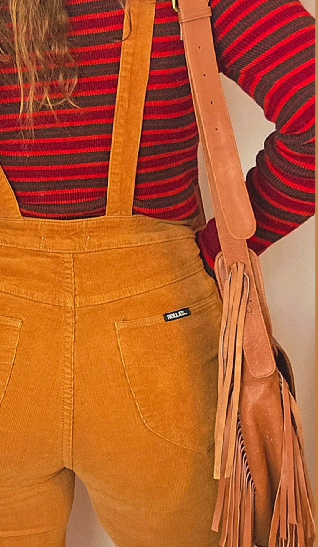 Person wearing mustard yellow overalls with a red and black striped shirt, holding a brown leather bag.