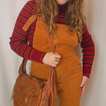 Person wearing mustard yellow overalls with a red and black striped shirt, holding a brown leather bag.