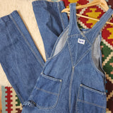 Blue denim overalls on a wooden hanger against a colorful geometric-patterned rug.