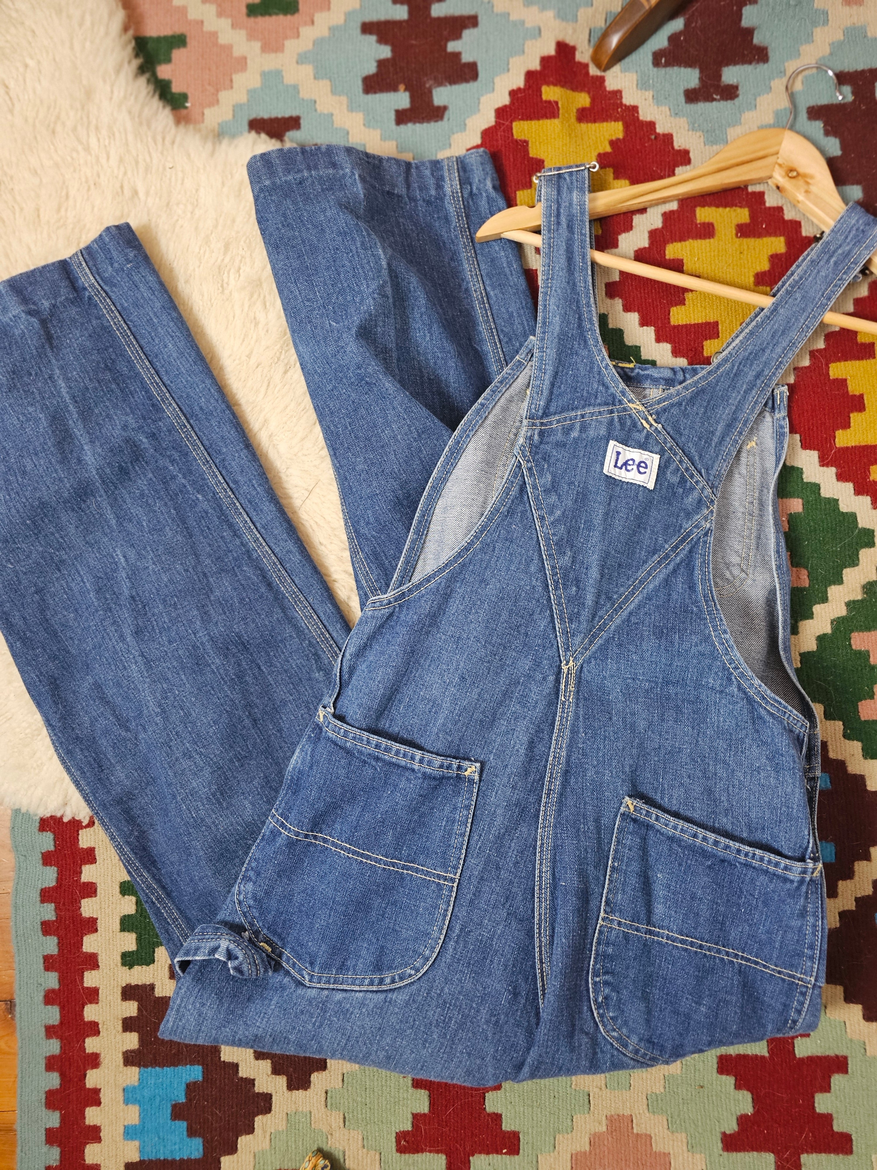 Blue denim overalls on a wooden hanger against a colorful geometric-patterned rug.