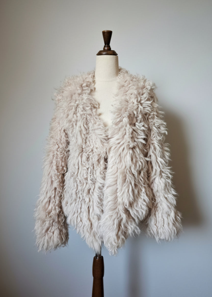 A shaggy, curly faux-shearling jacket in a light cream or oatmeal color, displayed on a cream-colored dress form mannequin against a neutral light gray wall.