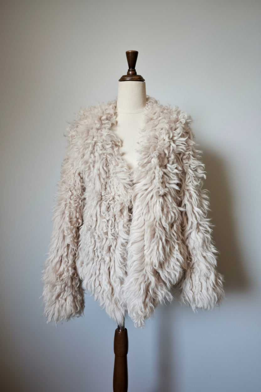 A shaggy, curly faux-shearling jacket in a light cream or oatmeal color, displayed on a cream-colored dress form mannequin against a neutral light gray wall.