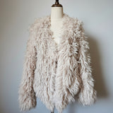 A shaggy, curly faux-shearling jacket in a light cream or oatmeal color, displayed on a cream-colored dress form mannequin against a neutral light gray wall.