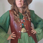 Person wearing a green dress, brown leather vest, and hat against a plain background