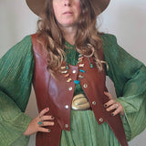 Person wearing a green dress, brown leather vest, and hat against a plain background