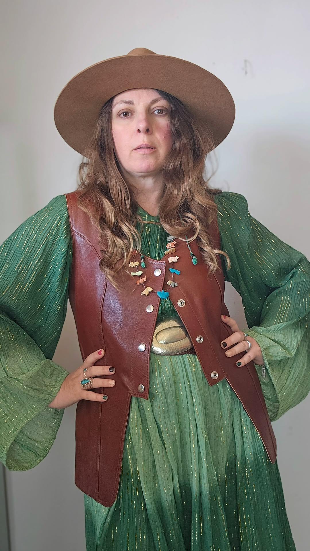 Person wearing a green dress, brown leather vest, and hat against a plain background