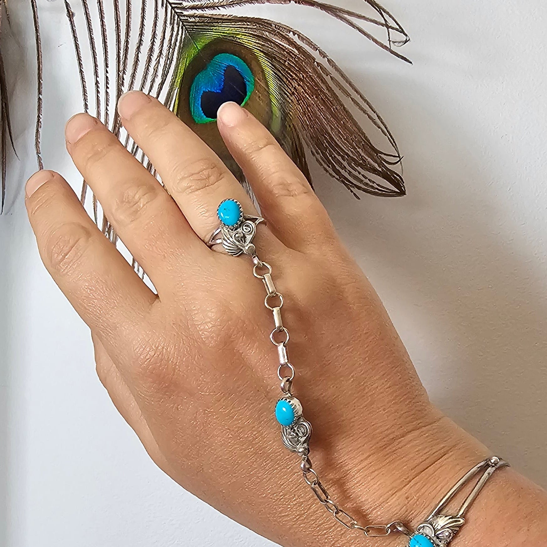Hand with turquoise ring and bracelet touching a peacock feather on a white background