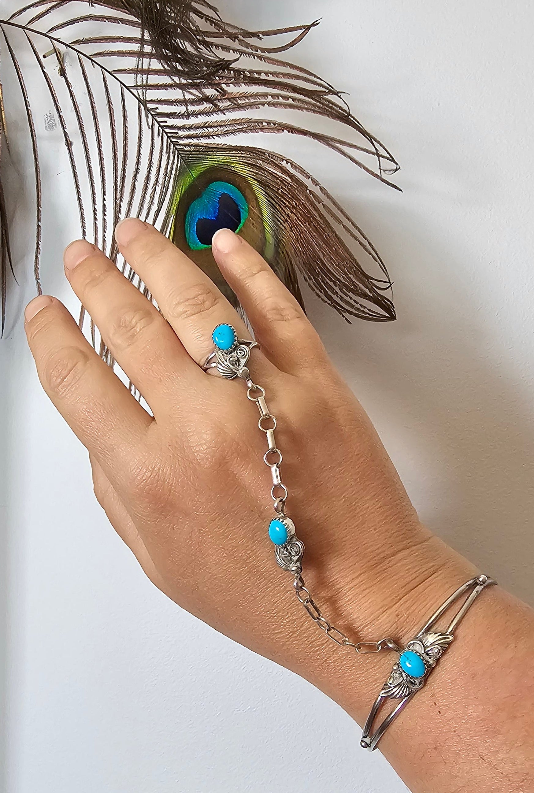 Hand with turquoise ring and bracelet touching a peacock feather on a white background