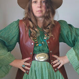 Person wearing a green dress with a brown vest and hat against a plain background