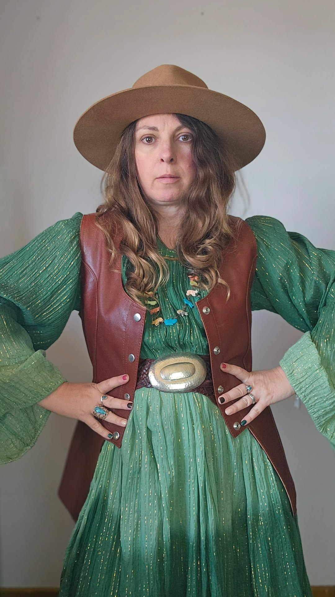 Person wearing a green dress with a brown vest and hat against a plain background