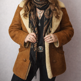 Woman wearing a brown coat with shearling lining, hat, and scarf against a plain background