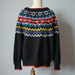 Black sweater with colorful pattern on a wooden hanger against a neutral wall.